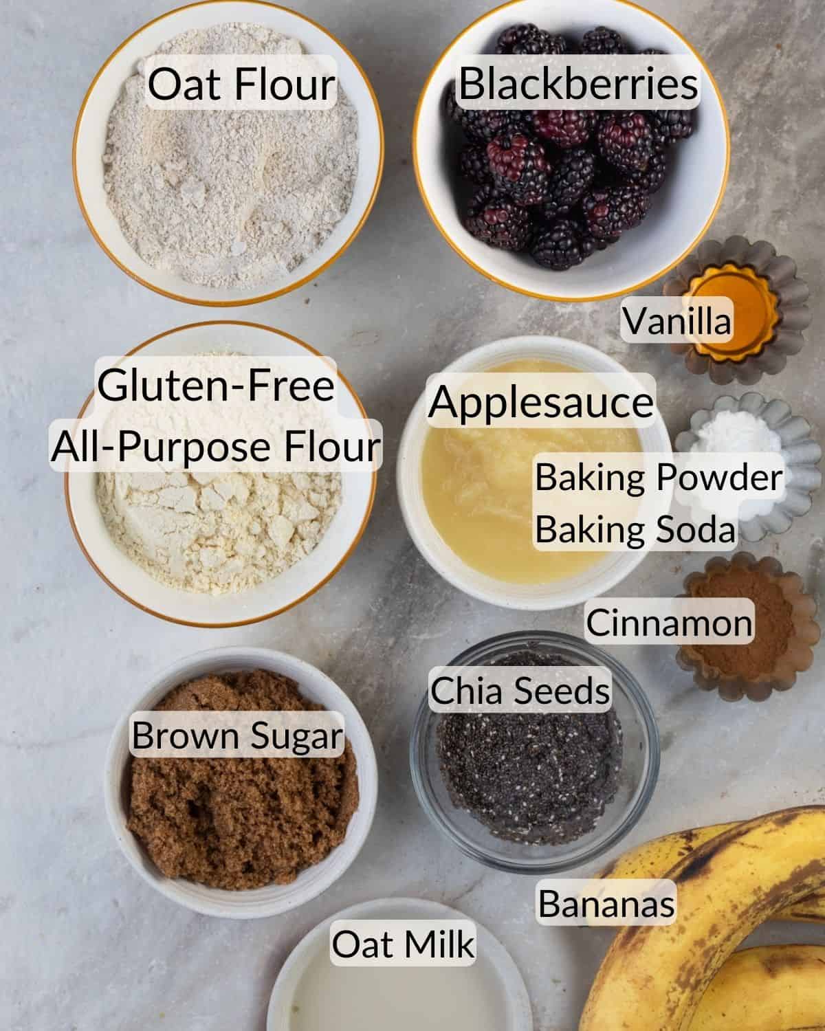 An assortment of baking ingredients neatly arranged on a marble countertop, labeled for preparing banana blackberry oatmeal muffins.