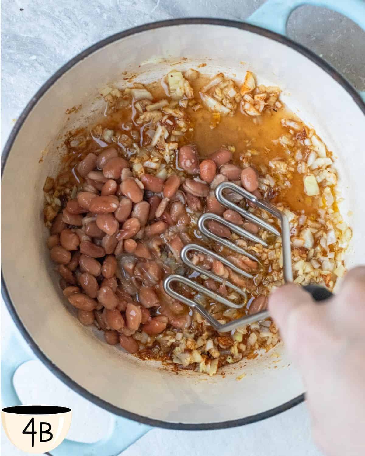 A potato masher in a pot with pinto beans and sautéed onions, crushing the beans to thicken the stew for a traditional Brazilian rice and beans texture.