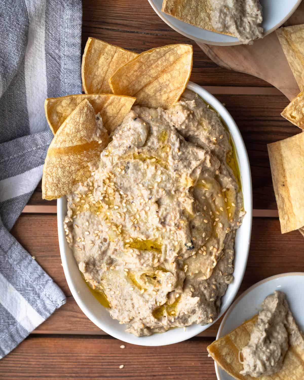 Overhead shot of eggplant tahini dip served in a white bowl, topped with sesame seeds and olive oil, with pita chips on the side, all set on a wooden surface.