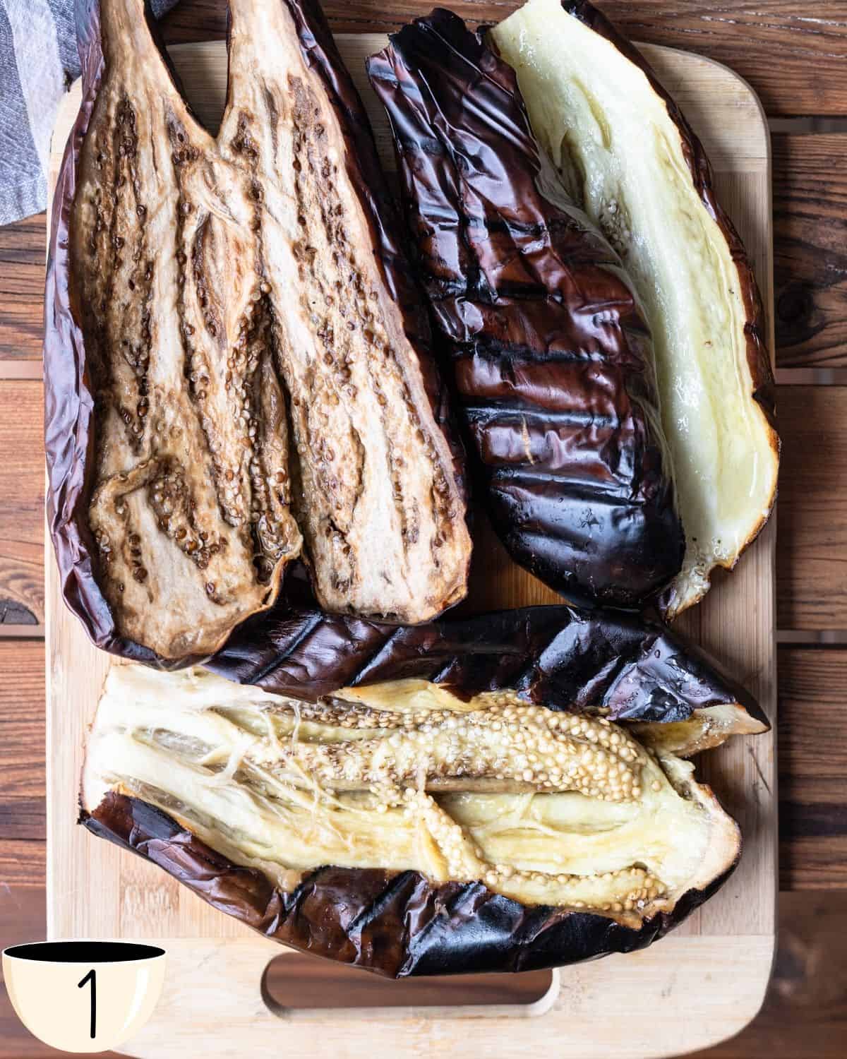 Roasted eggplants split open to reveal soft, cooked insides with a charred exterior, on a wooden cutting board, showing the preparation stage of a baba ganoush recipe