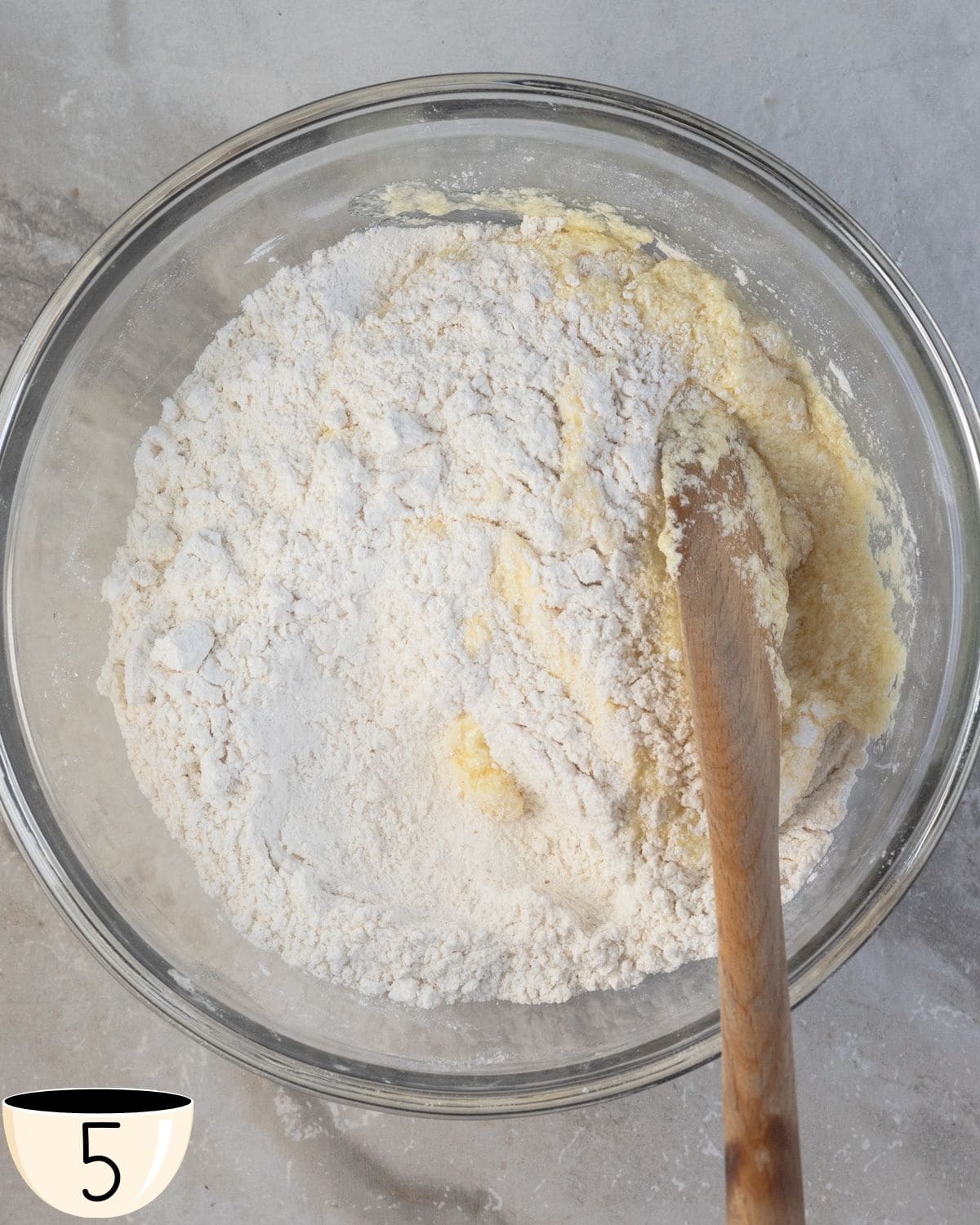 A large glass bowl with a mixture of gluten-free muffin batter and a wooden spoon, before the wet and dry ingredients are fully combined.