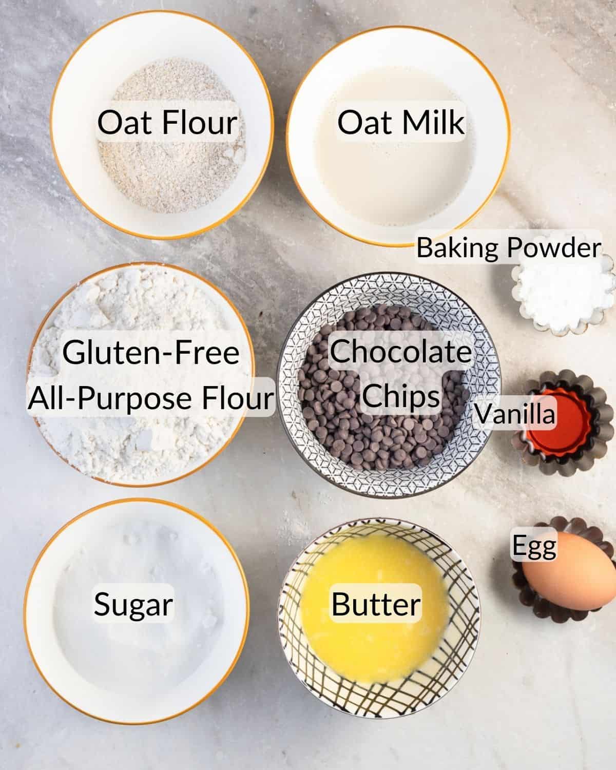 Ingredients for gluten-free chocolate chip muffins laid out on a marble surface, including bowls of oat flour, gluten-free all-purpose flour, chocolate chips, sugar, butter, an egg, vanilla extract, oat milk, and baking powder, with each ingredient labeled.