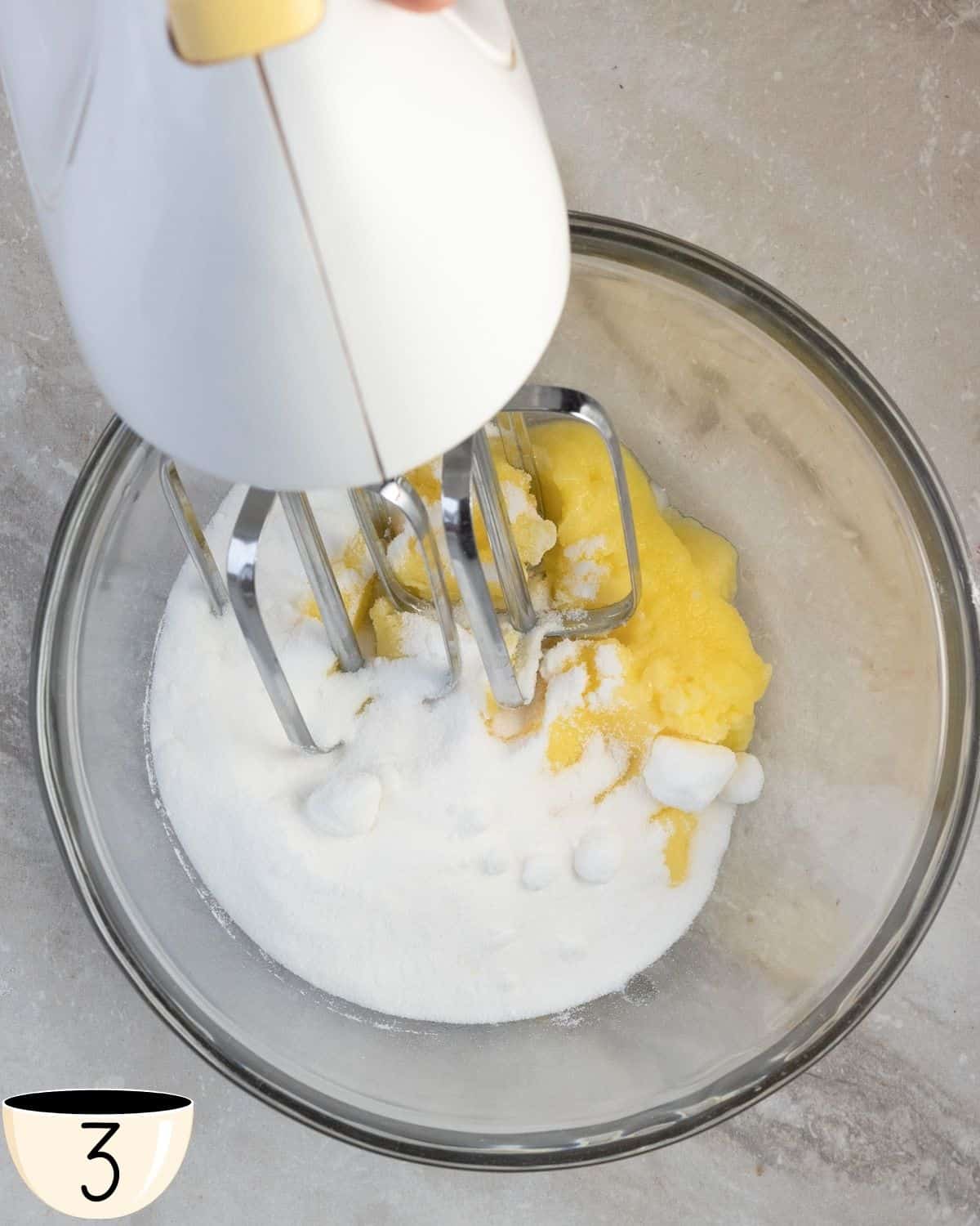 A hand mixer creaming together butter and sugar in a glass bowl, with a dusting of sugar around the bowl.