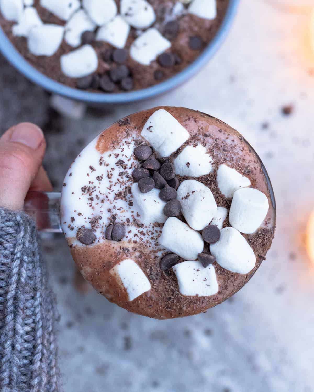 A top-down view of a mug of healthy hot chocolate, showing a generous topping of marshmallows and chocolate chips, held by hands in a snug sweater.