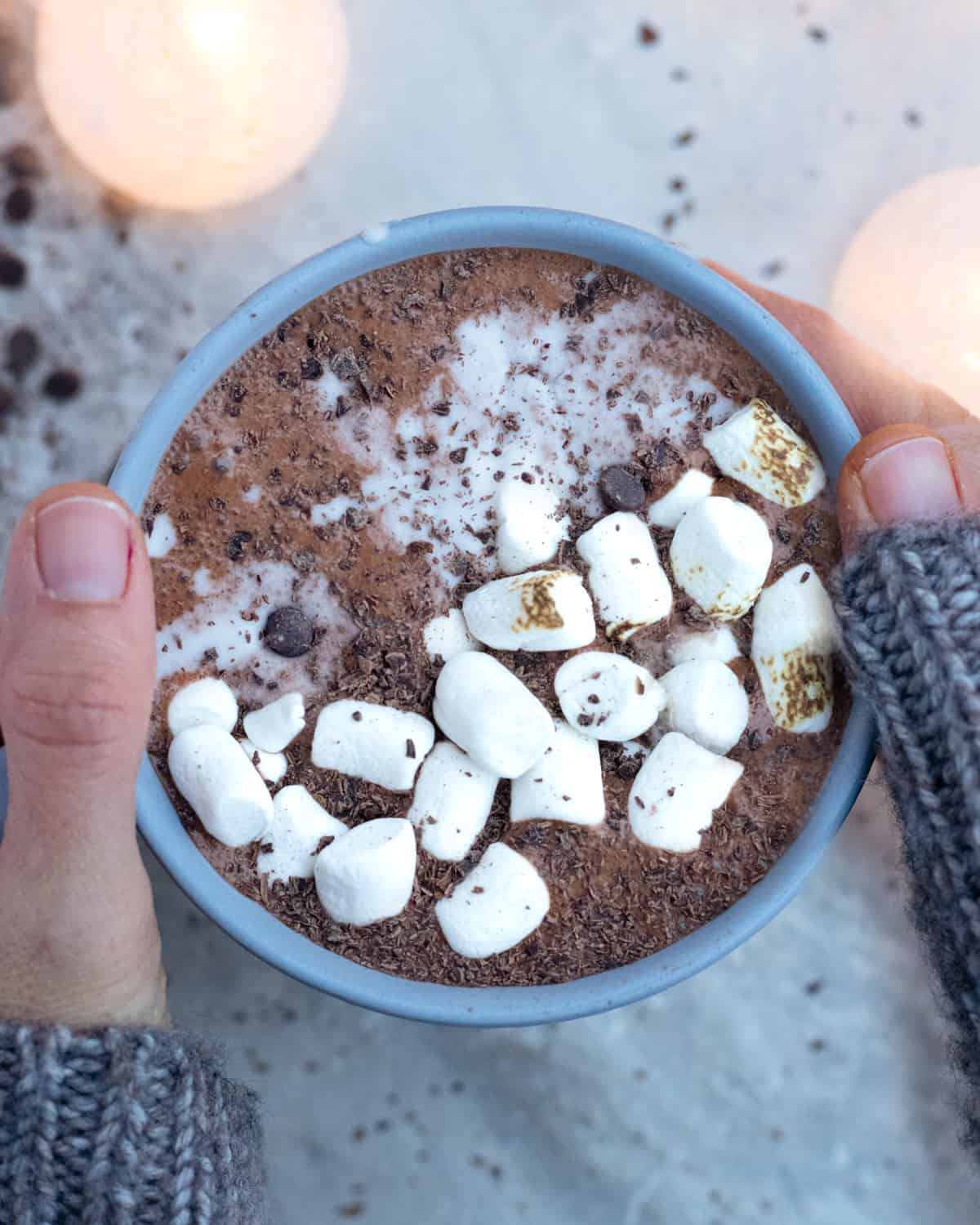 A cozy scene with hands wrapped in a knitted grey sweater holding a bowl of protein hot chocolate topped with marshmallows and a sprinkle of chocolate shavings, with soft glowing lights in the background.