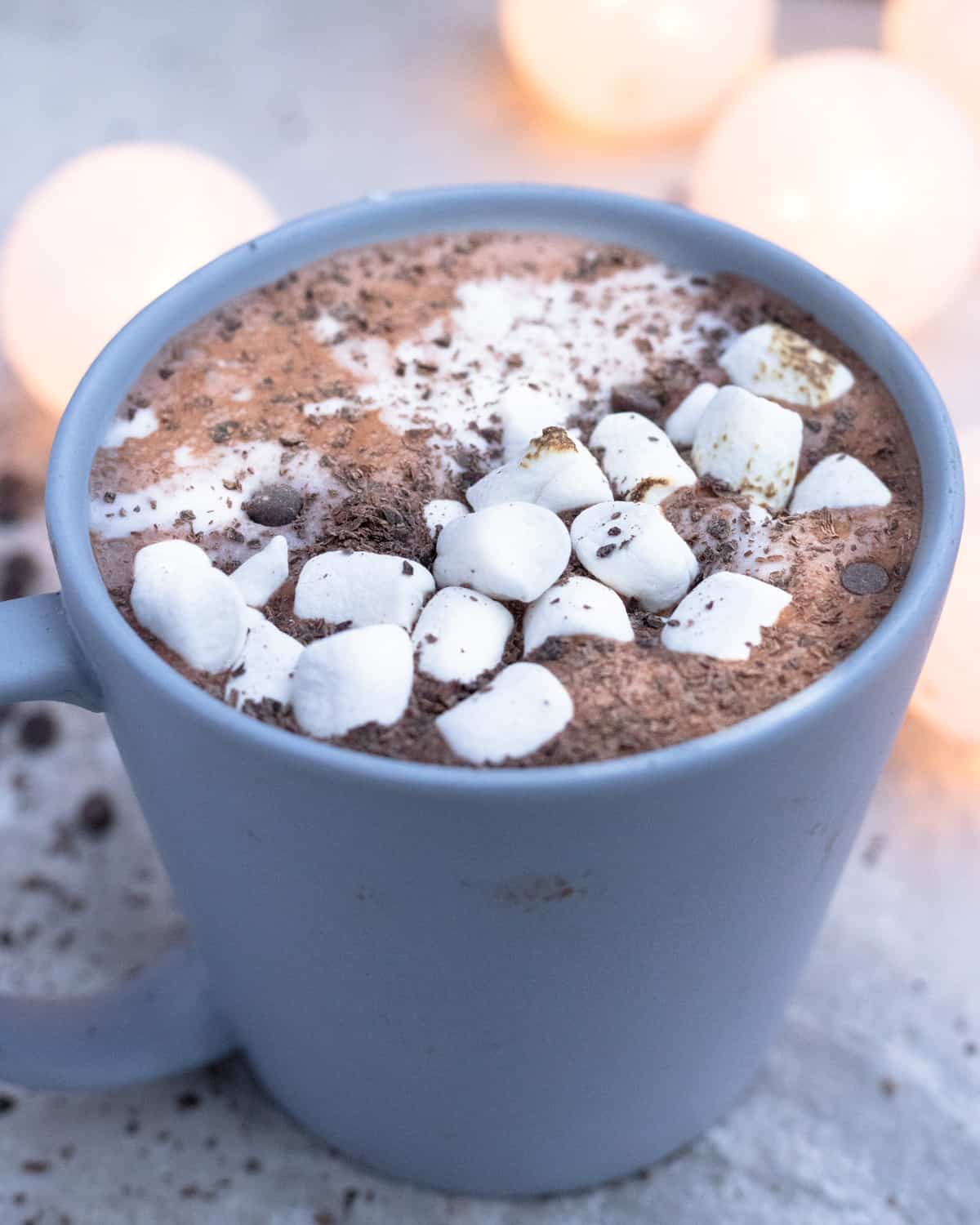A mug of protein hot chocolate richly topped with marshmallows and a dusting of cocoa powder, set against a backdrop of warm glowing lights.