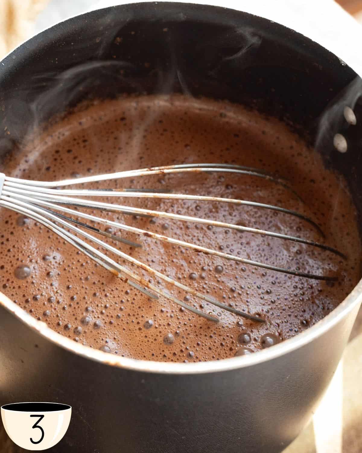 A pot on the stove with frothy hot chocolate with protein being whisked, demonstrating the cooking process of the beverage.