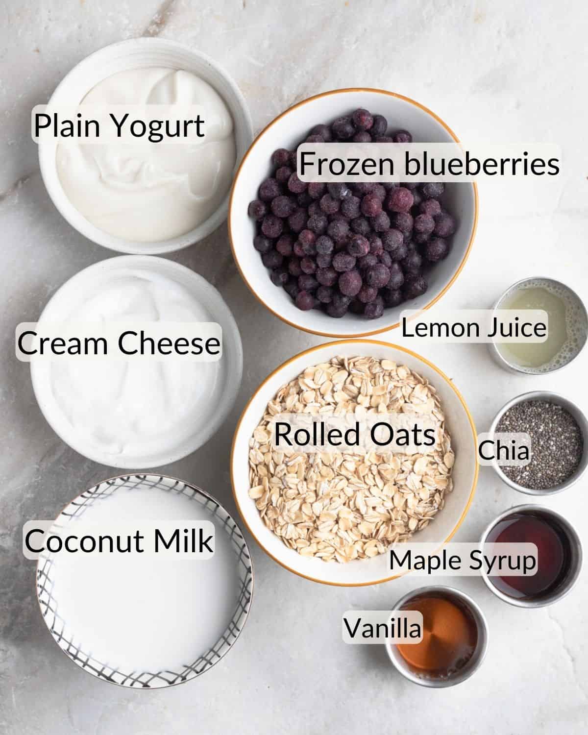 Ingredients for berry cheesecake overnight oats neatly arranged on a marble surface, including bowls of plain yogurt, frozen blueberries, cream cheese, rolled oats, chia seeds, coconut milk, maple syrup, lemon juice, and vanilla.