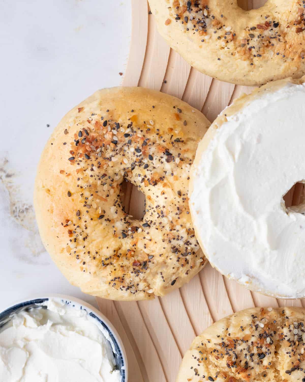 An overhead view of gluten-free bagels with Everything Bagel seasoning on a bamboo mat, next to a spreader and bowl of cream cheese.
