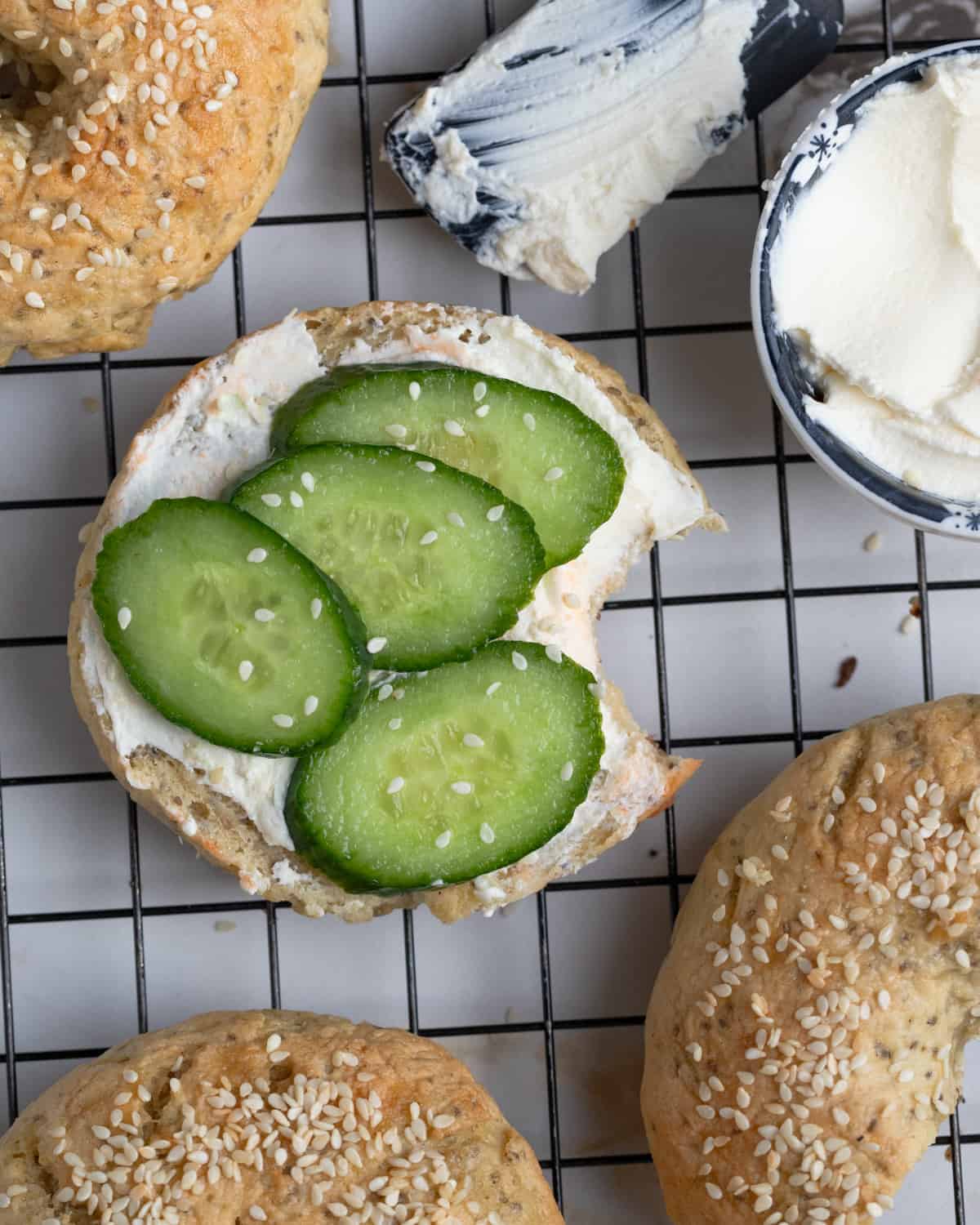 OVerhead of some vegan bagels with on esliced in half topped with vegan cream cheese and sliced cucumbers. The bagels are sitting on a wire rack.