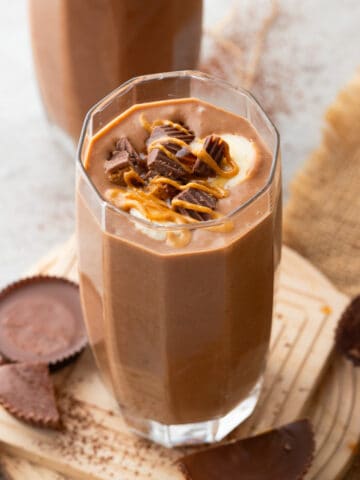 A peanut butter cup smoothie in a tall clear glass is topped with chopped peanut butter cups and a drizzle of peanut butter. It's placed on a wooden coaster with more peanut butter cups and a beige cloth in the background.