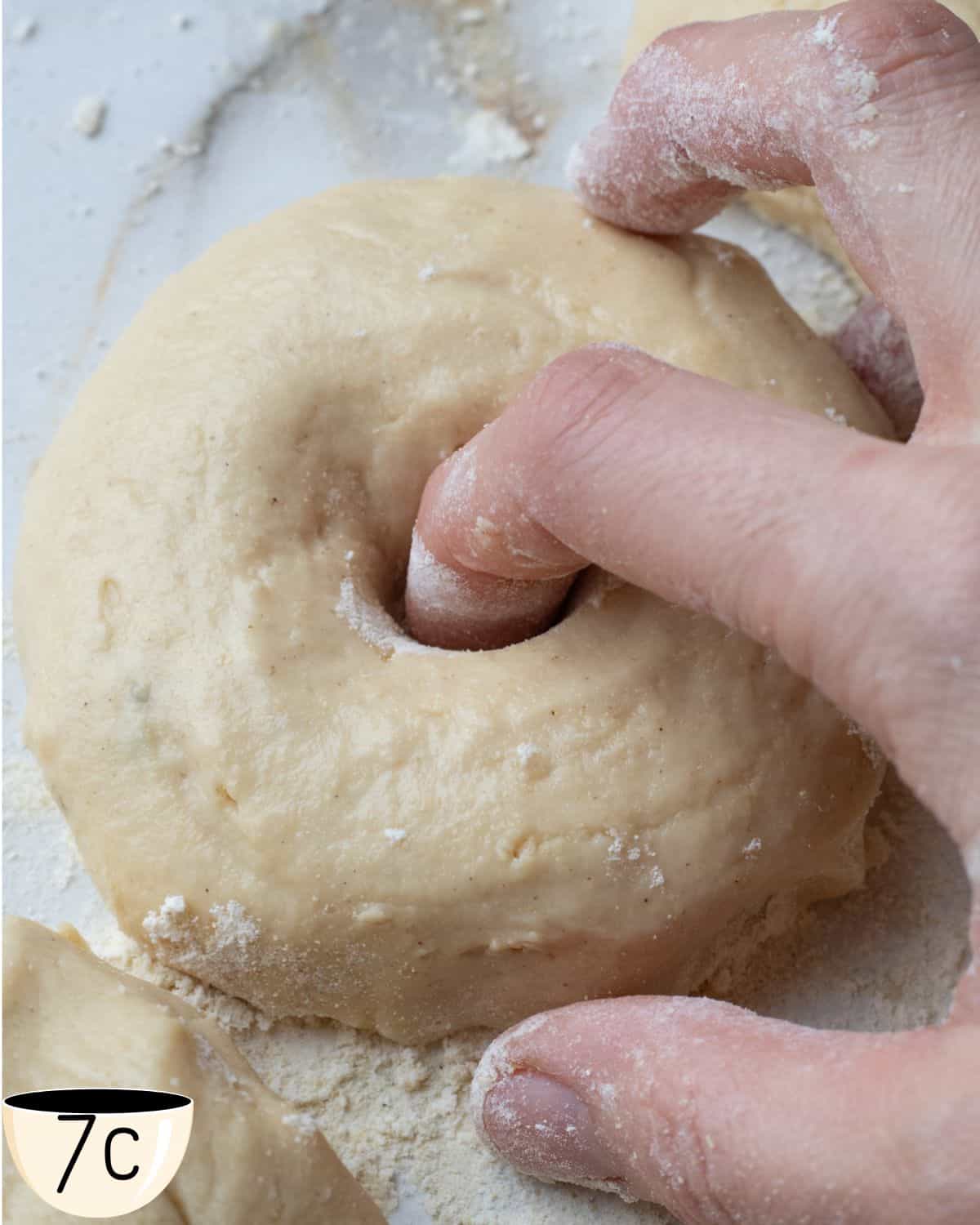 Fingers shaping a hole in the center of a gluten-free bagel dough ball on a floured surface.