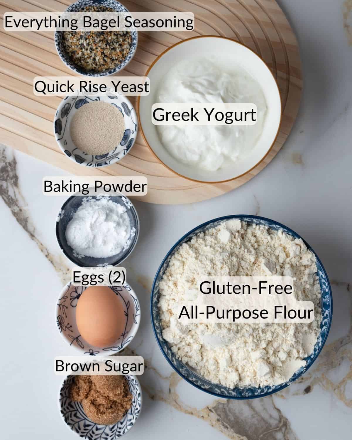 An array of ingredients on a bamboo mat for gluten-free bagels, including bowls labeled with Everything Bagel Seasoning, Quick Rise Yeast, Greek Yogurt, Baking Powder, two Eggs, Brown Sugar, and a larger bowl of Gluten-Free All-Purpose Flour.