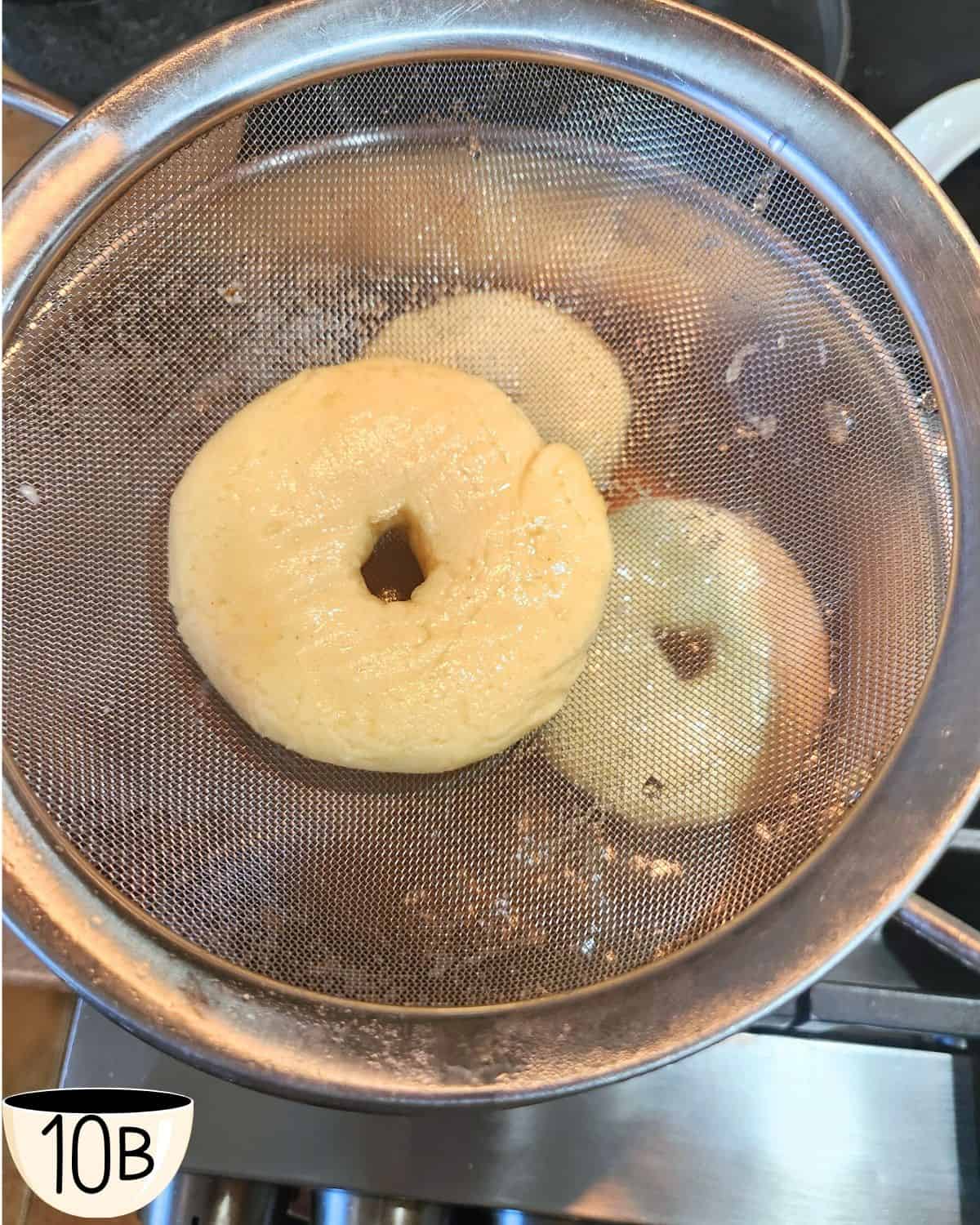 Three bagel being boiled in sugar water with one bagel being removed with a fine mesh sieve.