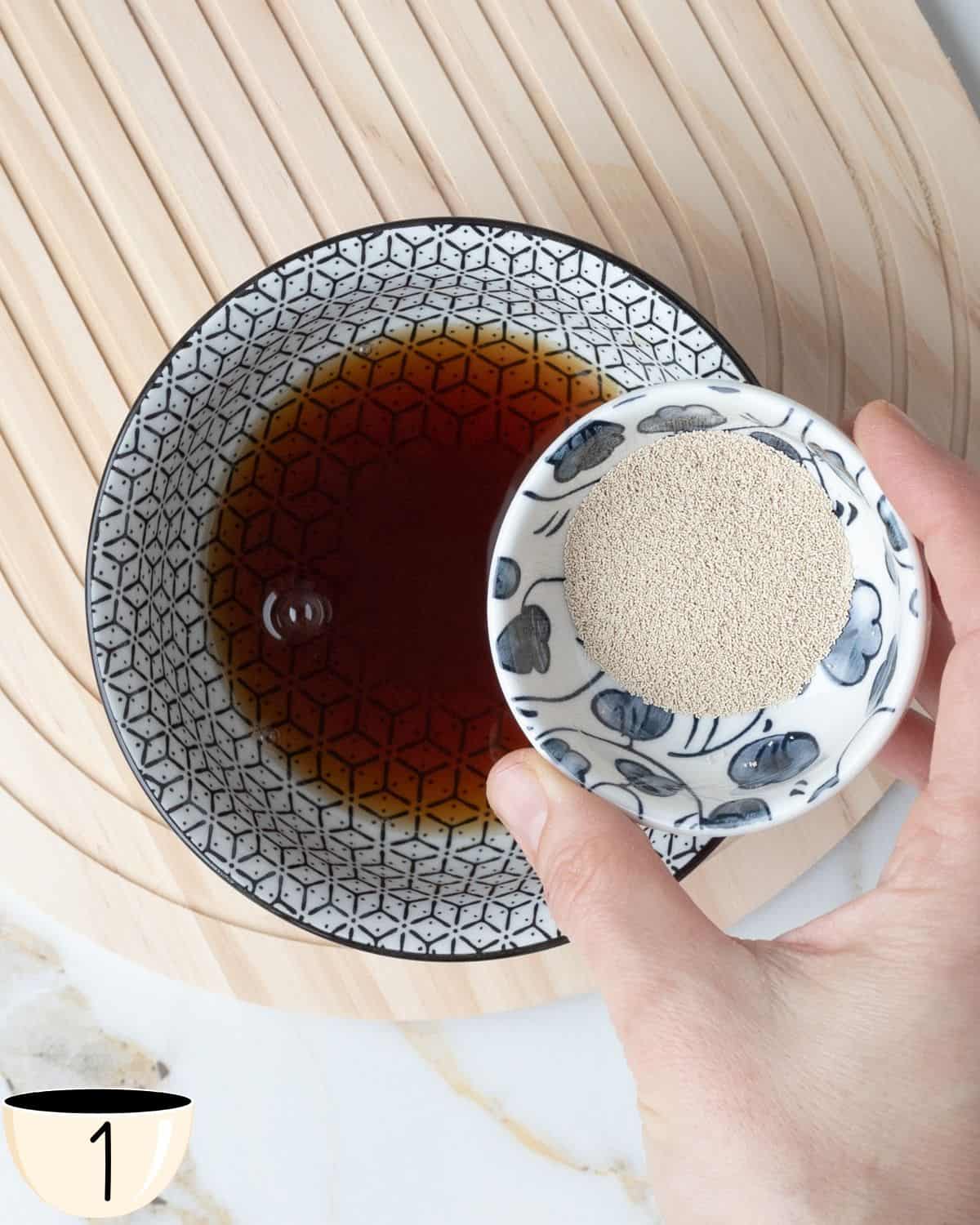A person's hand holding a small bowl with Quick Rise Yeast over a larger bowl containing water abd brown sugar to activate the yeast.