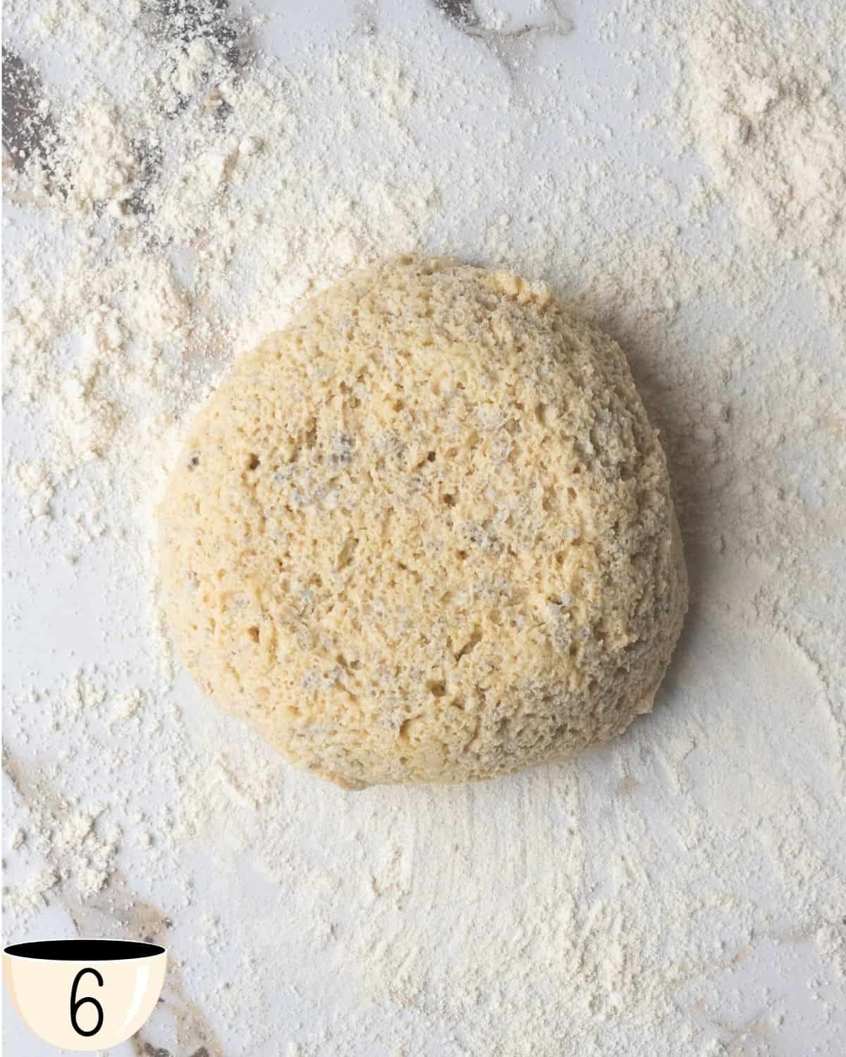 A single lump of gluten-free vegan bagel dough on a marble surface dusted with flour.