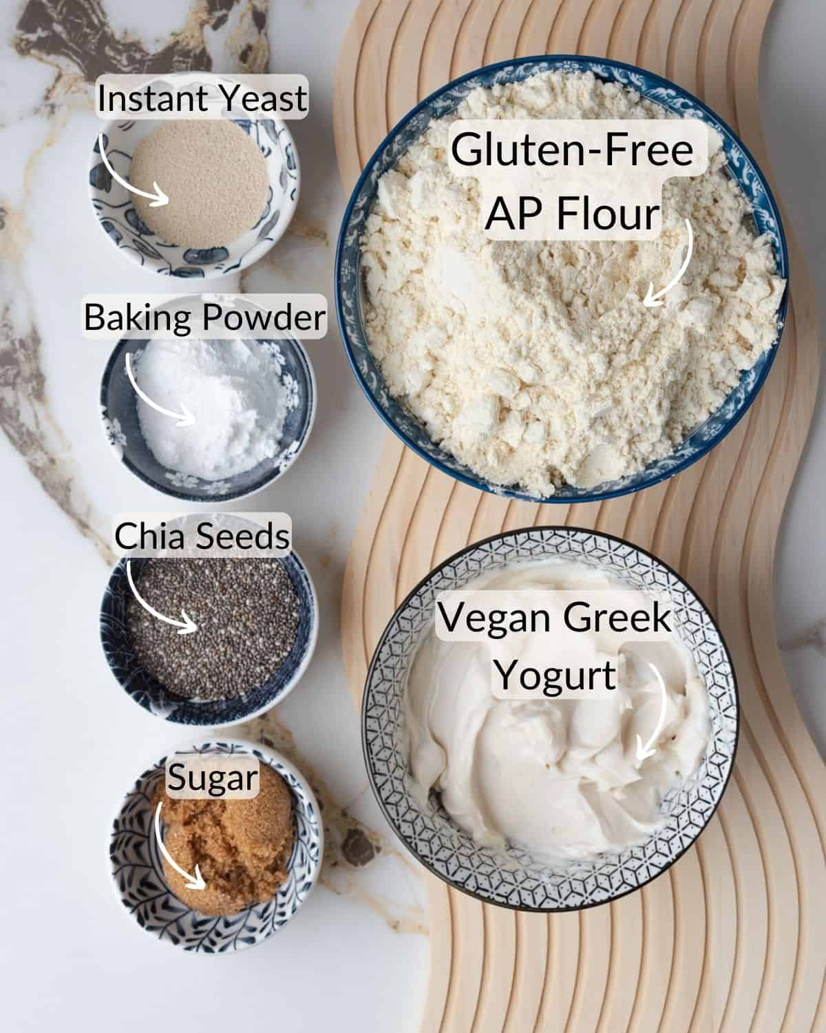 Ingredients for gluten-free vegan bagels laid out on a marble countertop, including bowls of instant yeast, baking powder, gluten-free all-purpose flour, chia seeds, sugar, and vegan Greek yogurt.