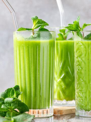 Three detox island green smoothies with a clear glass straw garnished with mint leaves. They are placed on a light, textured surface, with fresh mint leaves scattered around, offering a refreshing and healthy vibe and showing thie creamy texture and vibrant green color of the smoothie recipe.
