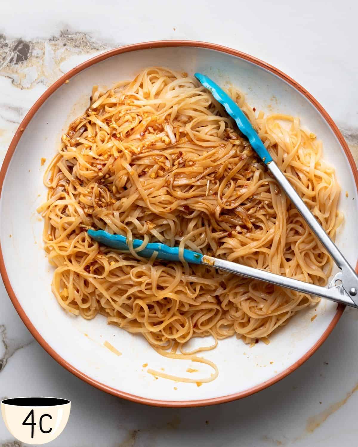 A bowl of seasoned noodles with a pair of tongs on a marble countertop.