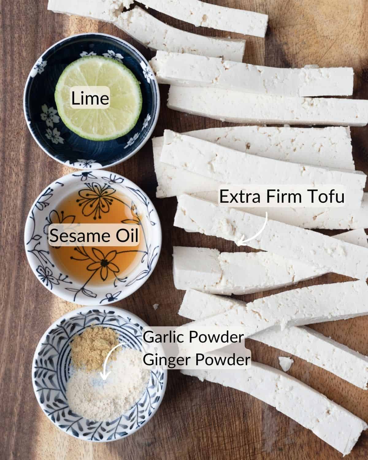 Ingredients for a spicy spring roll recipe on a wooden surface, featuring extra firm tofu strips, a bowl of sesame oil, and a bowl with a lime wedge.