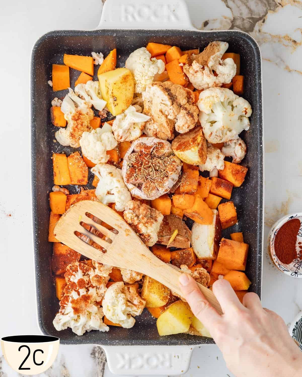 A wooden spatula mixing seasoned cauliflower and pumpkin pieces on a baking tray to ensure even coating before roasting.