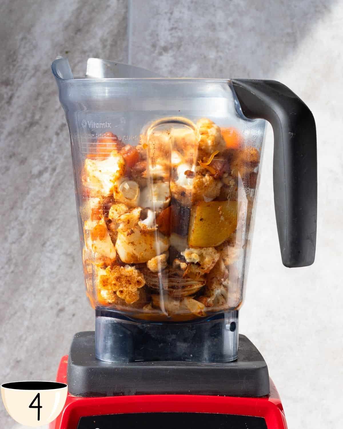 A blender half-filled with roasted vegetables including orange pumpkin and white cauliflower pieces, set against a kitchen backdrop.