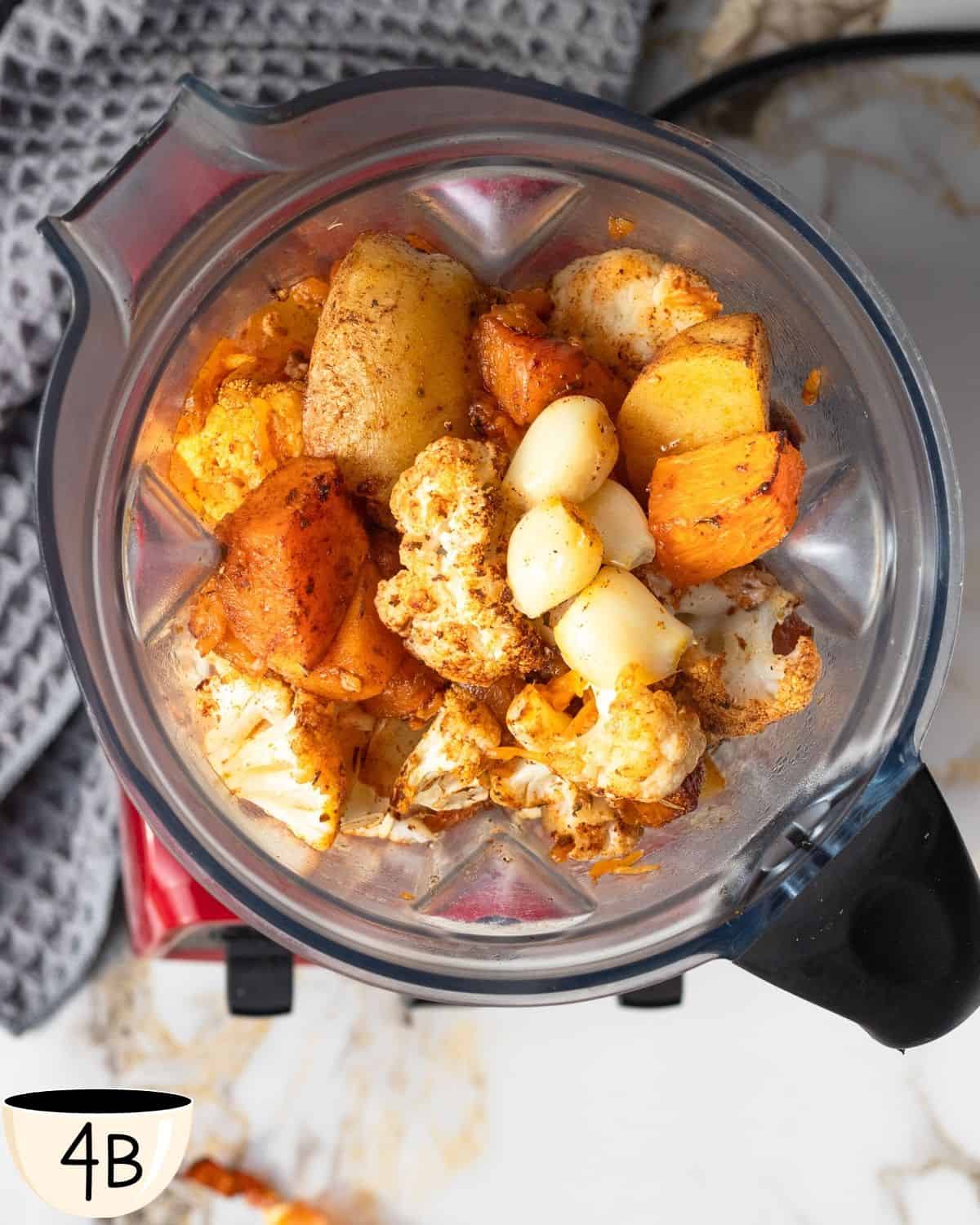 Looking down into a blender, we see roasted cauliflower and pumpkin with a wooden spoon, poised to blend into soup.