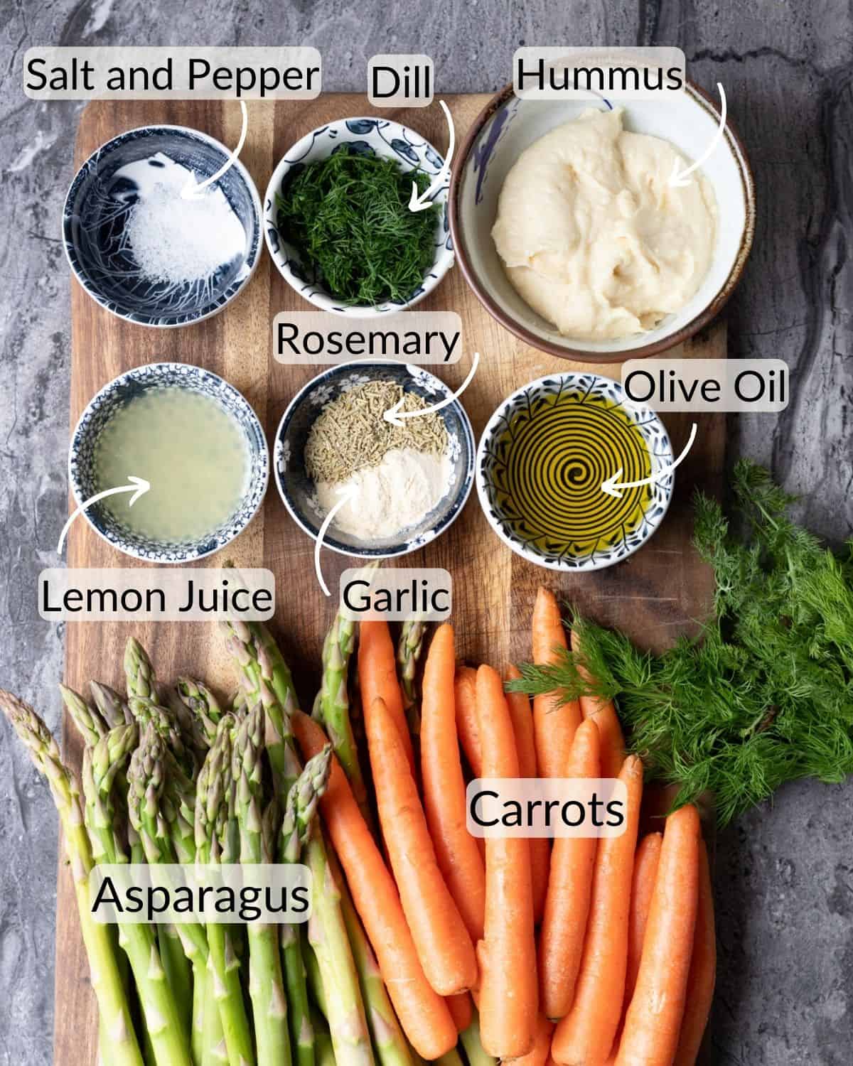 A top-down view of a wooden cutting board displaying labeled bowls of ingredients for a roasted veggetable recipe, including salt and pepper, dill, hummus, rosemary, garlic, olive oil, and lemon juice, alongside fresh asparagus and carrots.