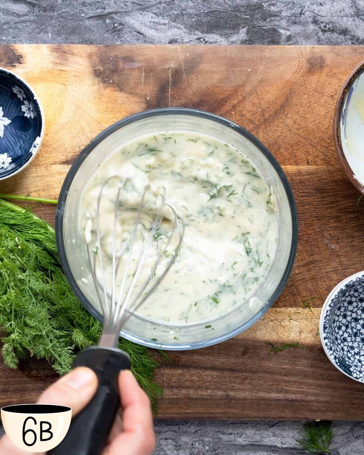 A hand whisking a creamy herb sauce in a glass bowl set on a wooden cutting board, with fresh dill and small bowls containing various ingredients around it.