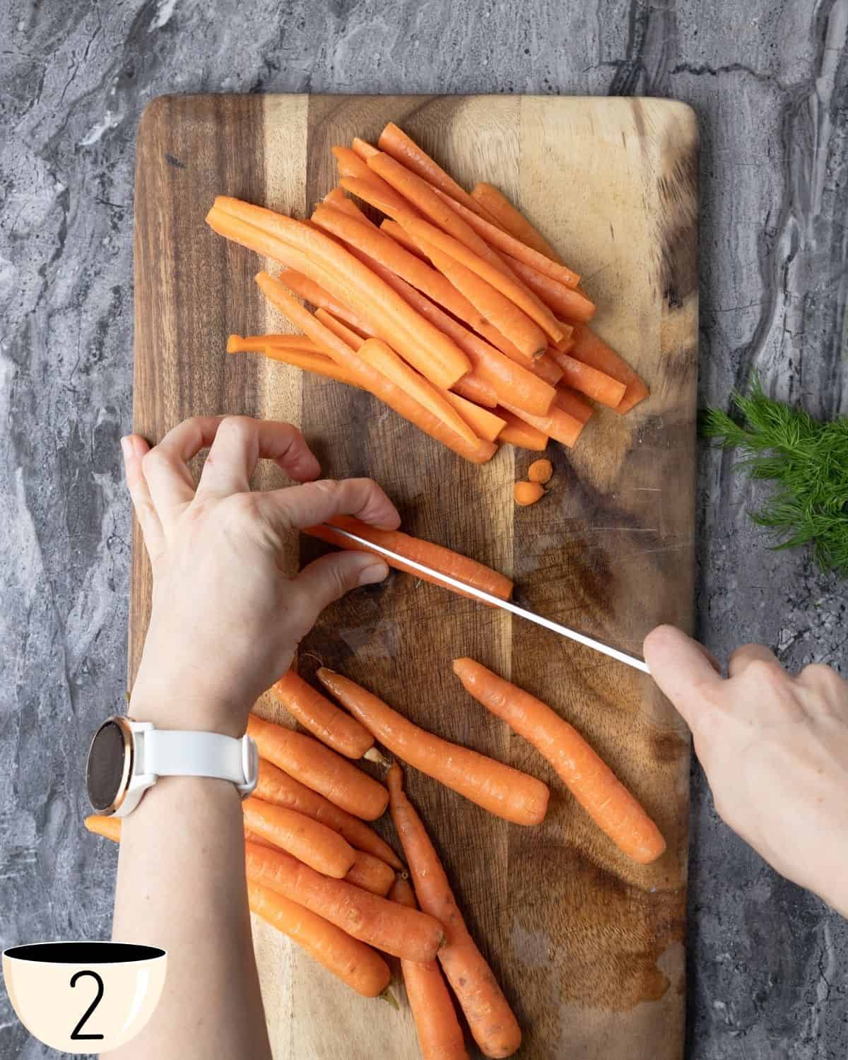A person's hands peeling and cutting carrots on a wooden cutting board, with a small bowl and fresh dill in the background.