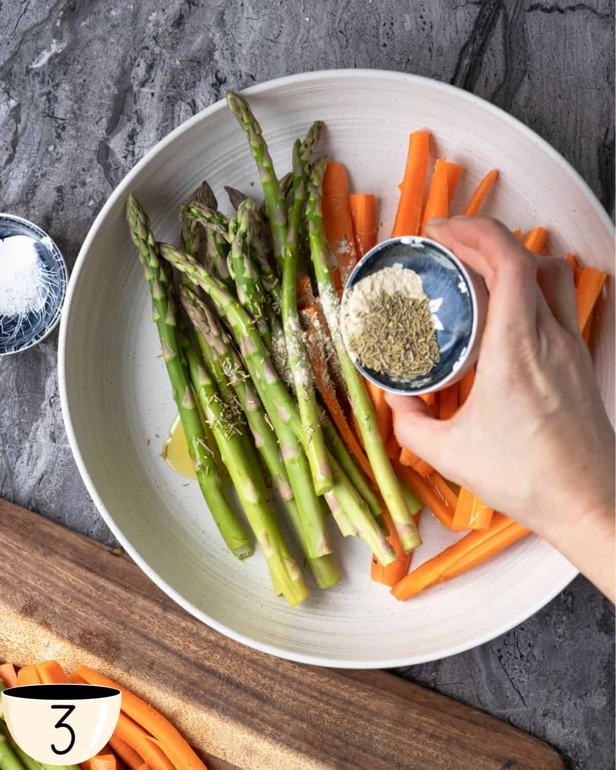 A person's hand sprinkling herbs over asparagus and carrots in a large white bowl on a wooden cutting board, with a bowl of garlic powder in the foreground.