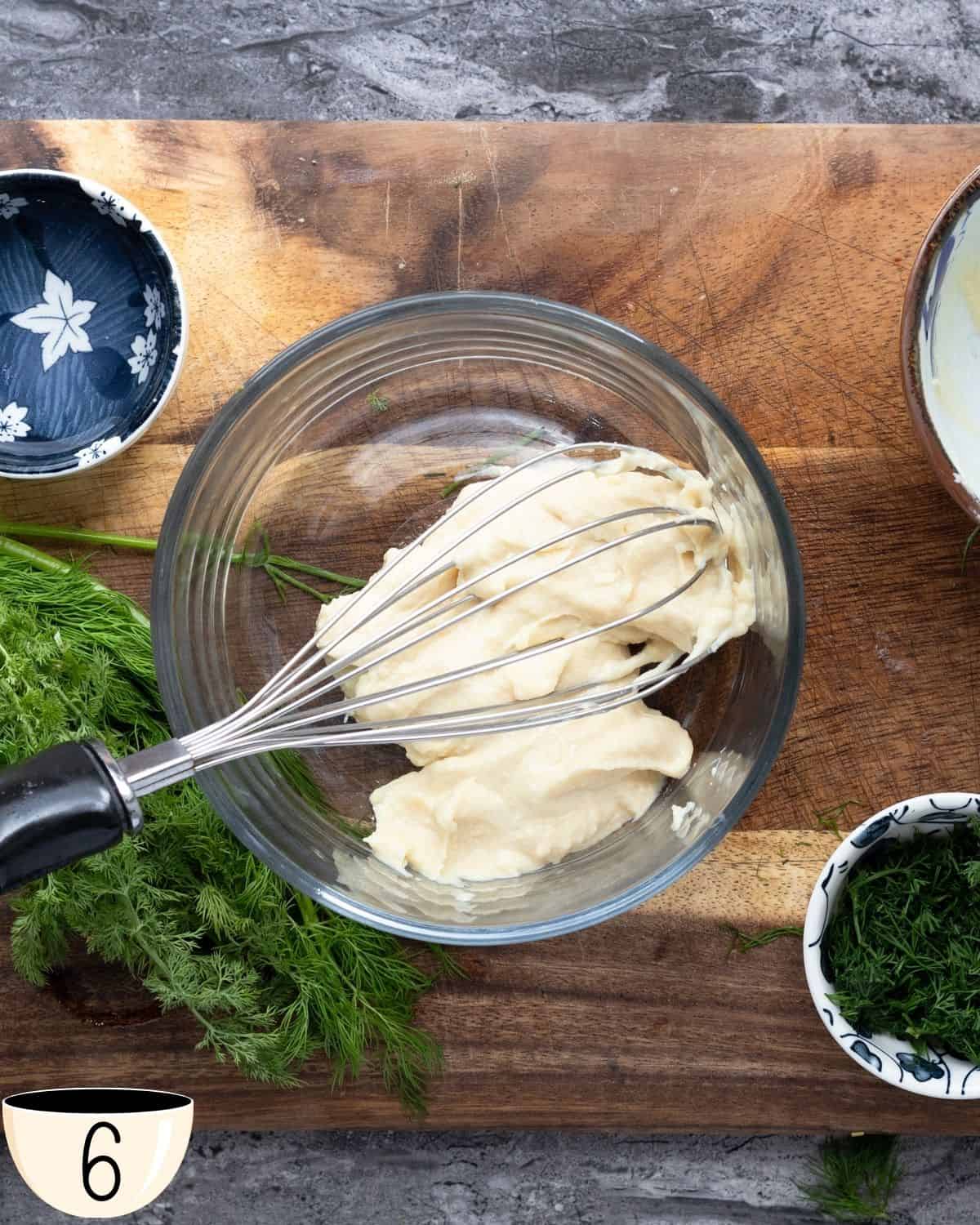 A glass bowl containing a thick, creamy sauce made with hummus, set on a wooden cutting board with fresh dill to the side.
