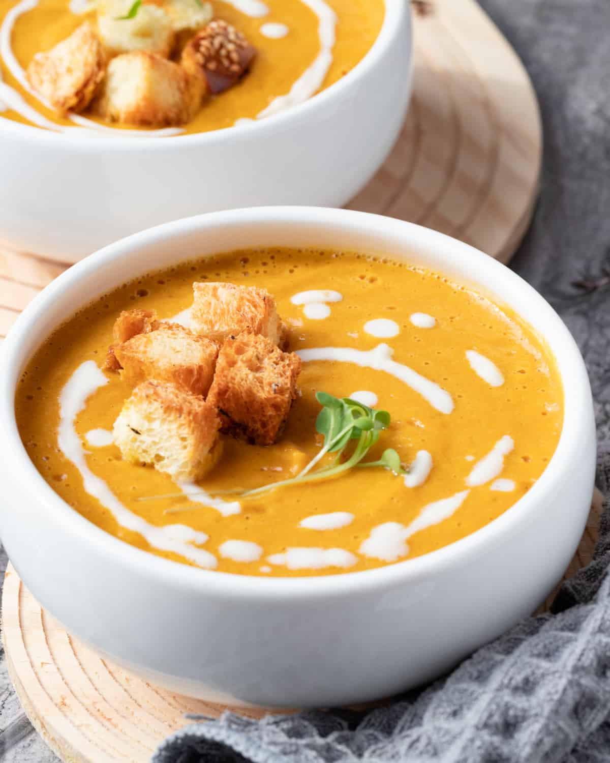 Close up of a bowl of creamy roasted pumpkin soup in a white bowl sitting on a wooden cutting board. The soup is granished with gluten-free croutons and a creamy drizzle.