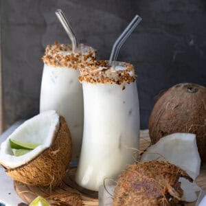 A close-up of two glasses of batida de coco with toasted coconut rims, garnished with toasted coconut flakes and lime wedges, surrounded by coconut halves and lime wedges.