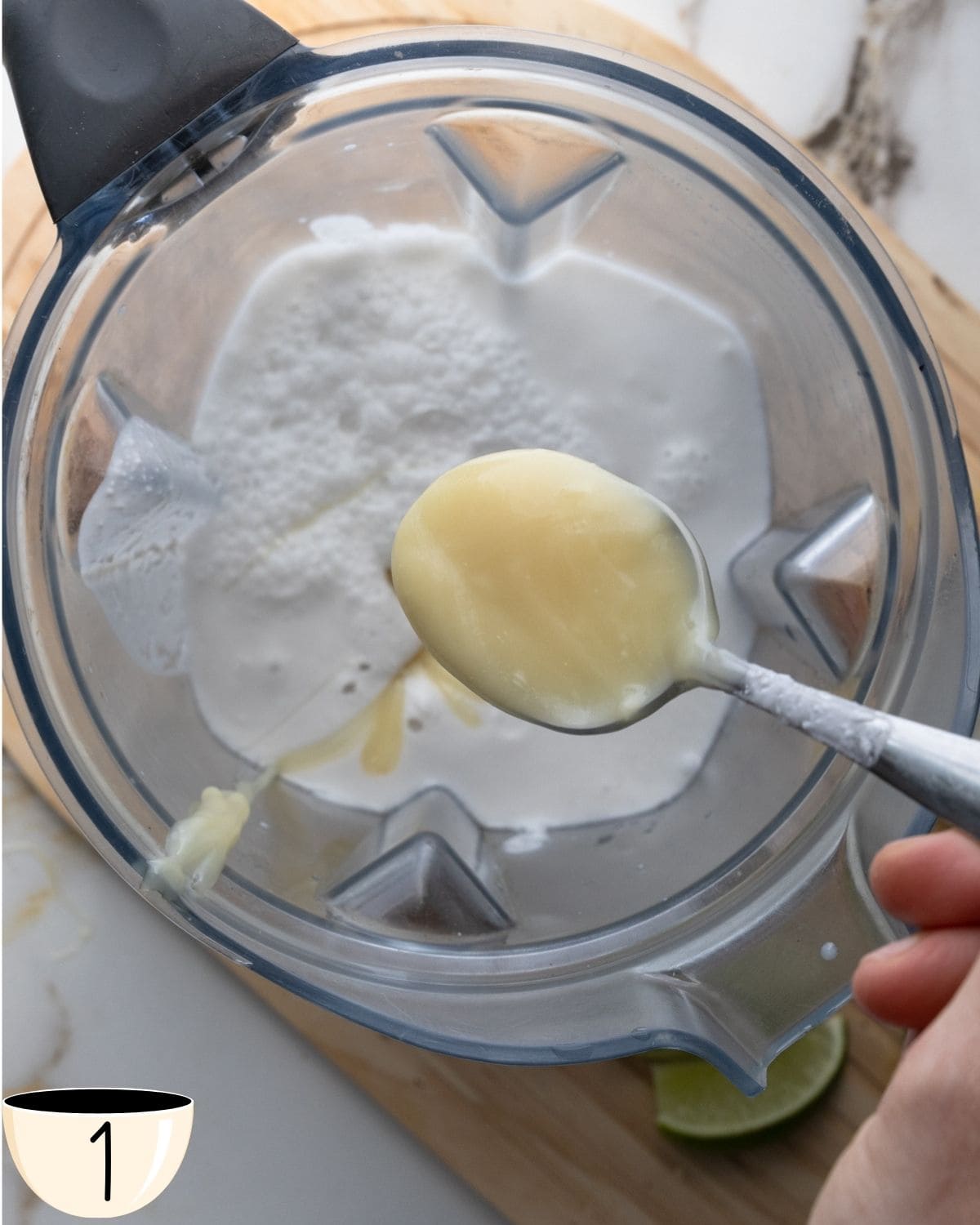 Close-up of a blender with coconut milk and a spoonful of condensed coconut milk being added.