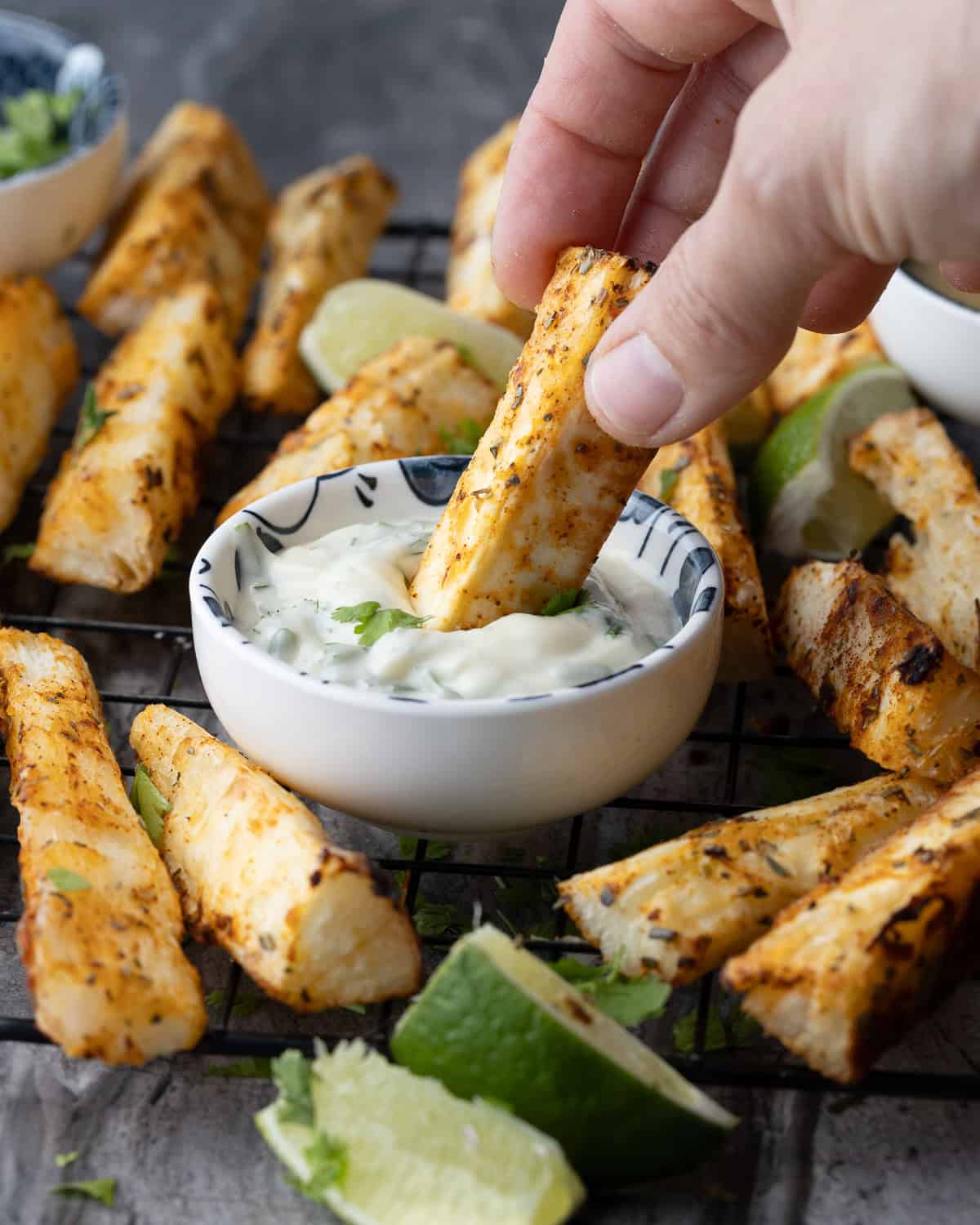 A hand dipping a crispy air fryer yuca fry into a bowl of creamy cilantro lime dipping sauce, with more yuca fries and lime wedges in the background.