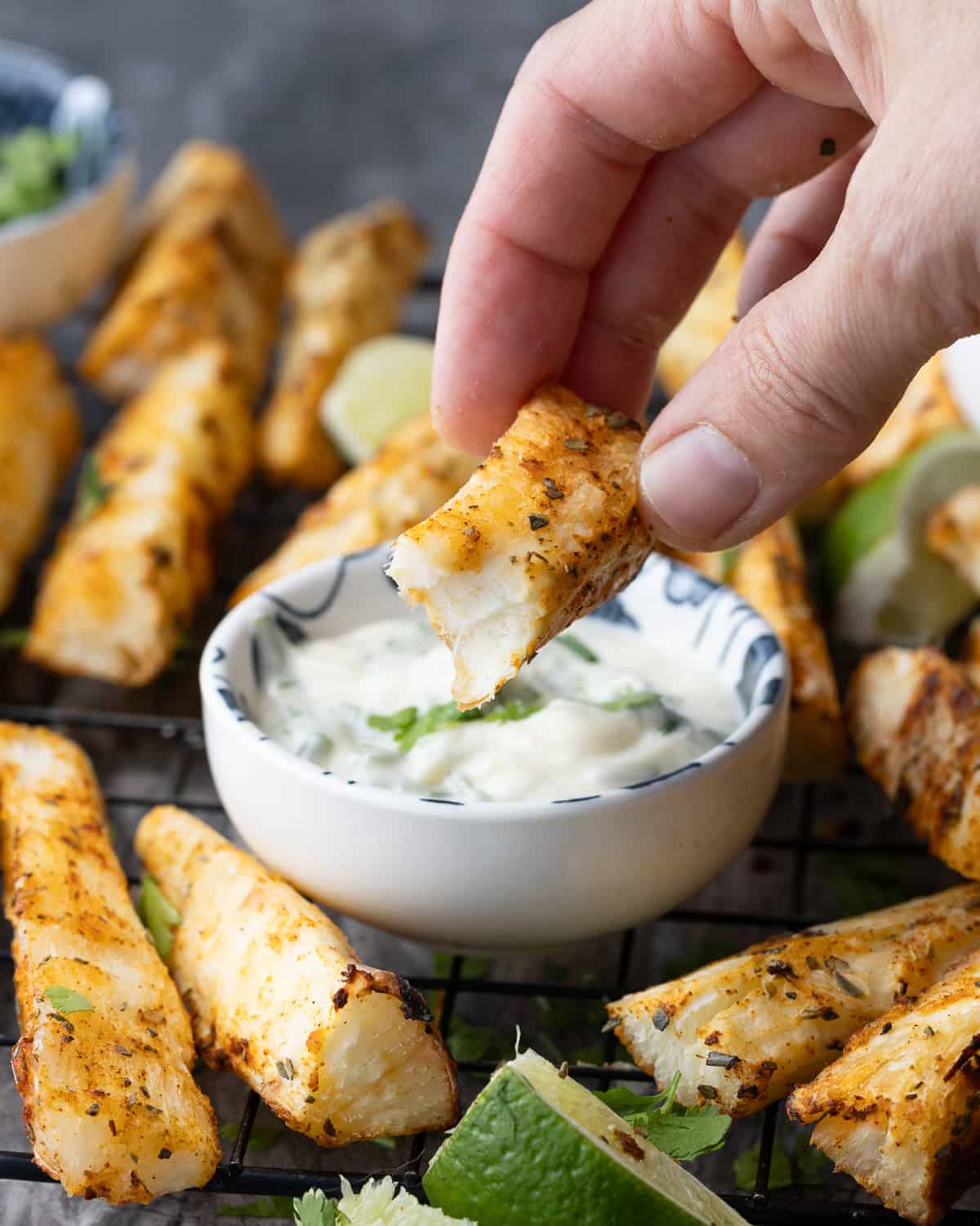 A hand holding a bitten yuca fry, showing the crispy exterior and fluffy interior, with a bowl of dipping sauce and more yuca fries and lime wedges in the background.