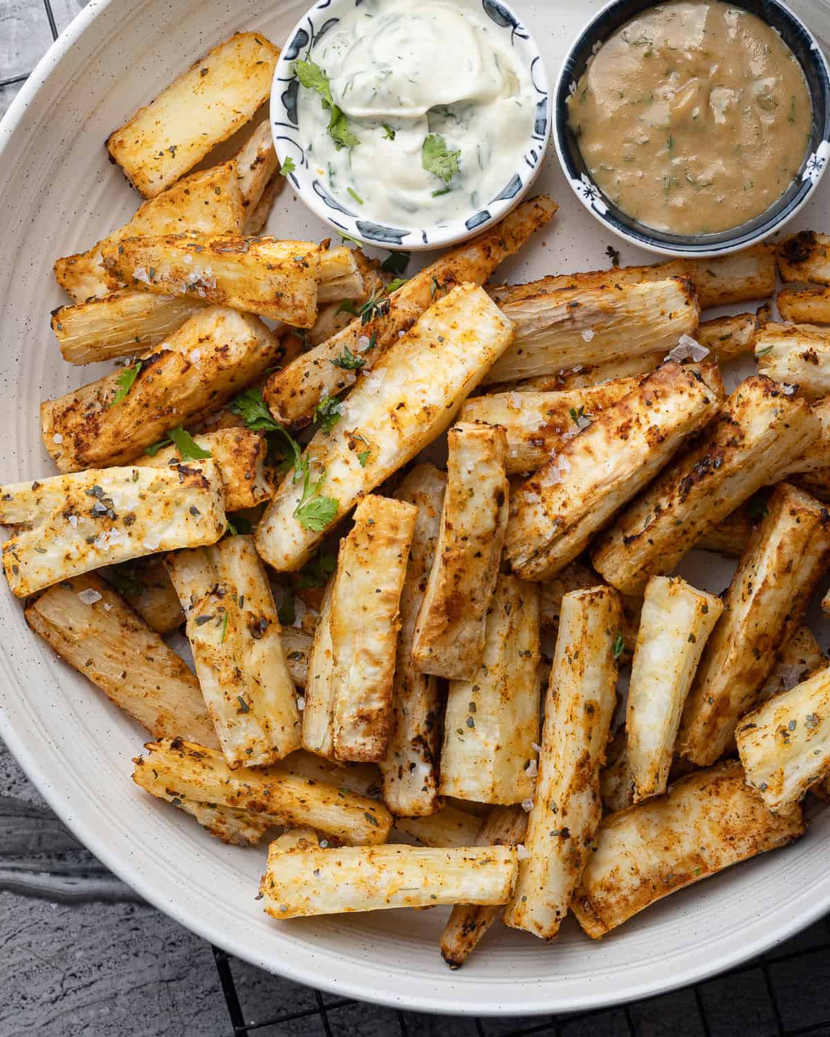 Overhead view of a large plate filled with air fried mandioca frita or yuca fries, garnished with cilantro and served with lime wedges and bowls of creamy dipping sauce.