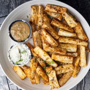 A bowl filled with crispy, seasoned yuca fries garnished with fresh cilantro and coarse sea salt, served alongside two small bowls of dipping sauces.
