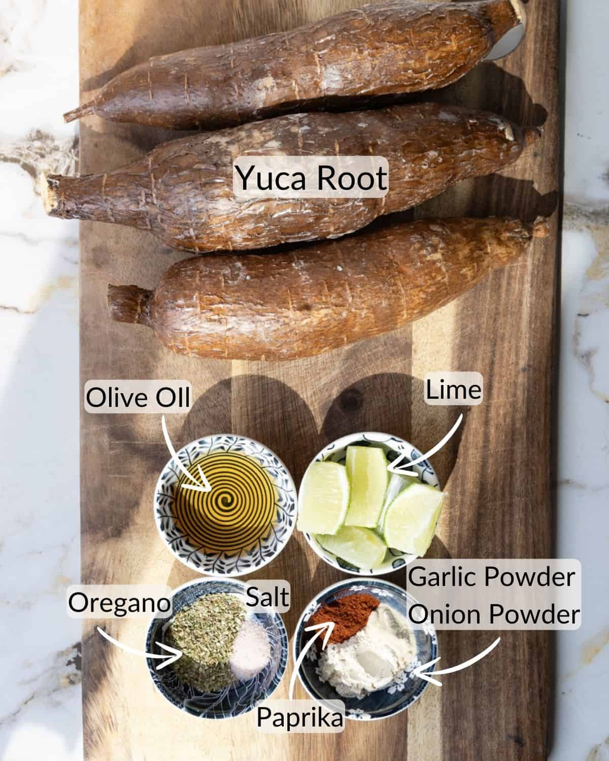 Three whole yuca roots, a bowl of olive oil, lime wedges, and small bowls containing oregano, salt, paprika, garlic powder, and onion powder displayed on a wooden cutting board.