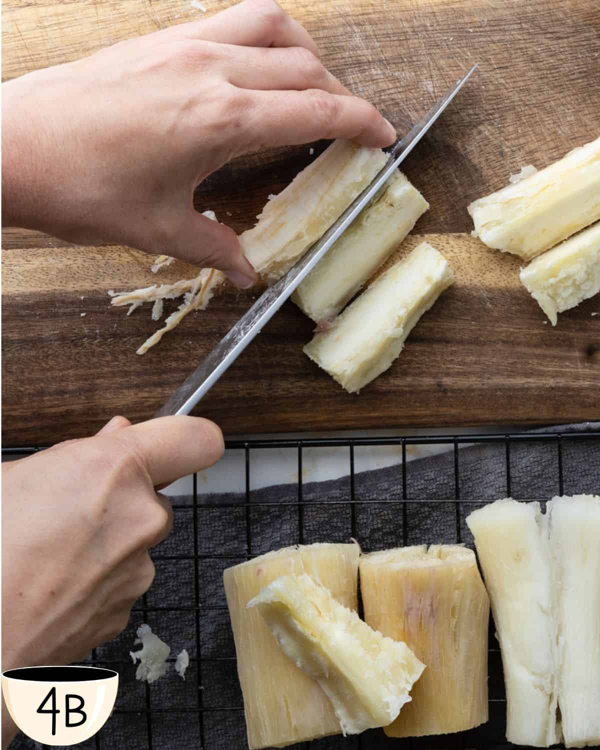 A hand using a sharp knife to cut boiled yuca into fry-sized pieces on a wooden cutting board, with more yuca pieces on a wire rack below.