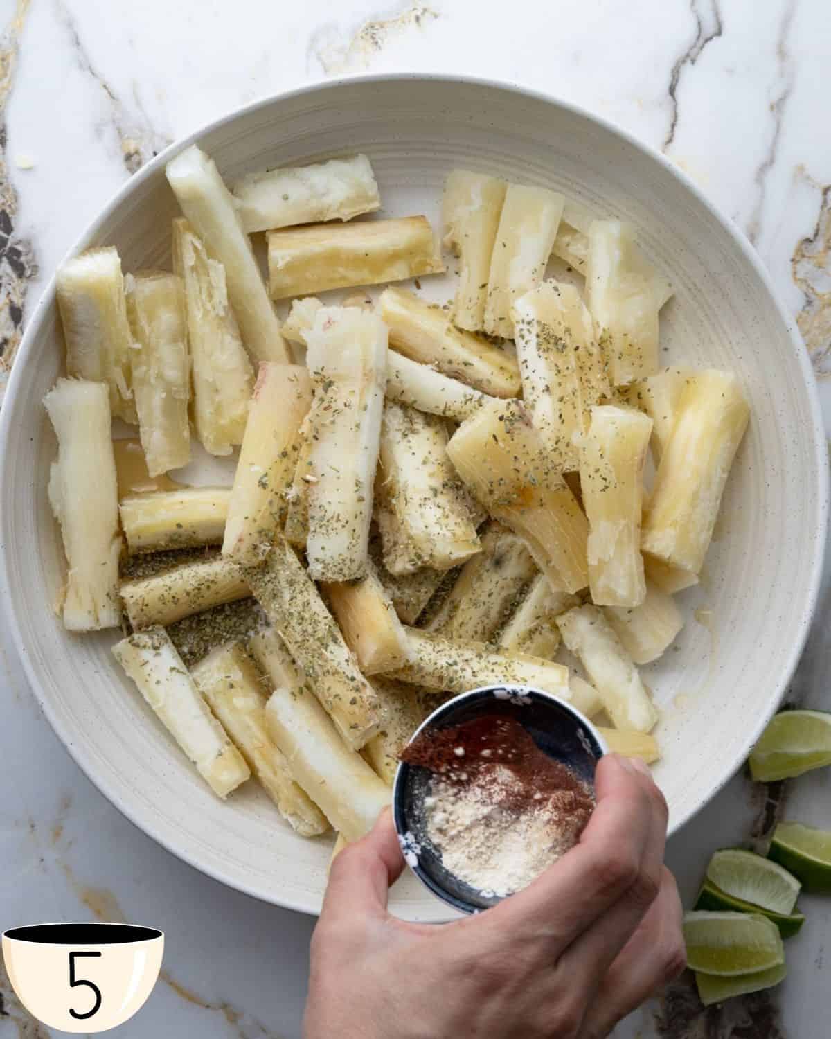 A hand sprinkling a spice mixture over yuca fries in a large white bowl, preparing to season them.
