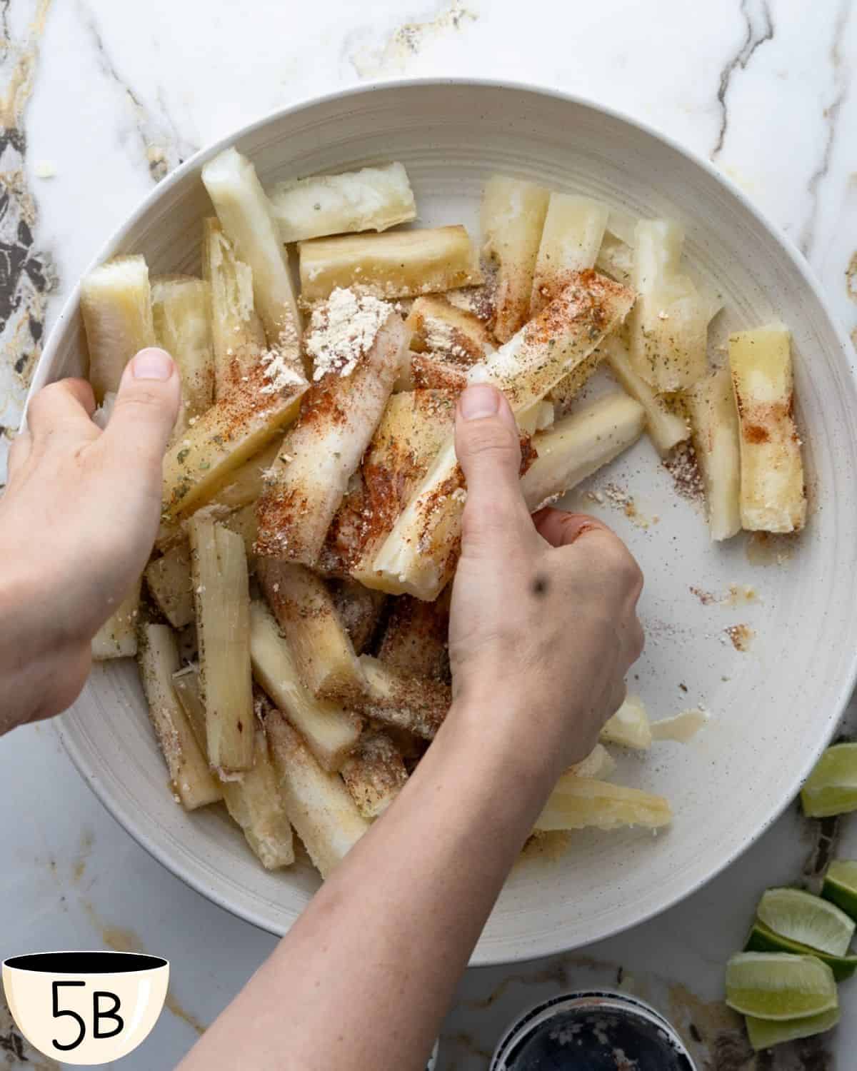 Two hands tossing yuca fries with seasoning in a large white bowl, ensuring even coating of the spices.