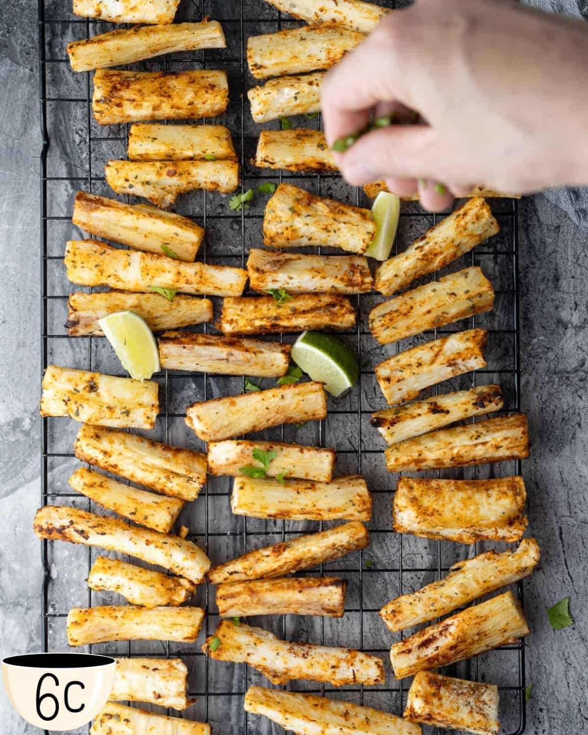 A hand sprinkling chopped cilantro over seasoned yuca fries on a wire rack, with lime wedges placed among the fries.