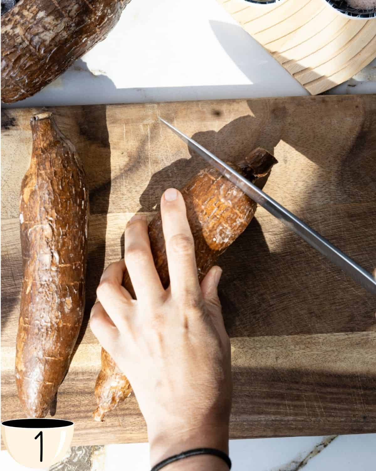 A hand holding a yuca root while using a sharp knife to cut off the ends on a wooden cutting board.