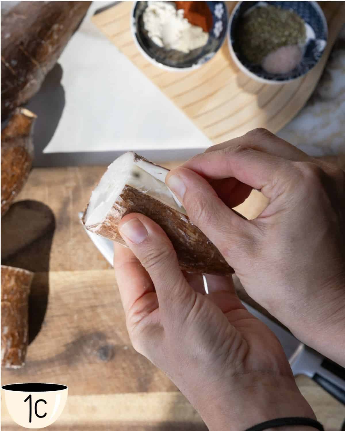 A hand peeling the outer thick skin off a piece of yuca root to reveal the white flesh inside.