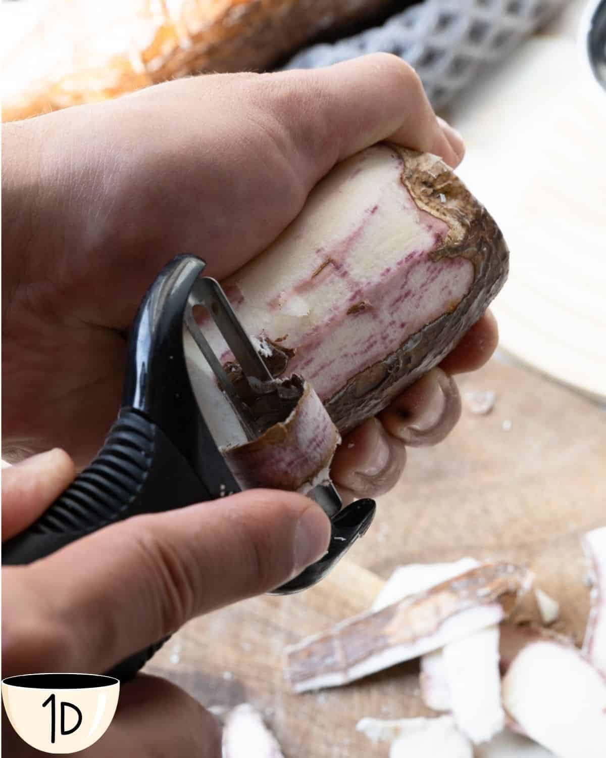 A hand using a vegetable peeler to remove the tough skin from a piece of yuca root, exposing the white flesh.