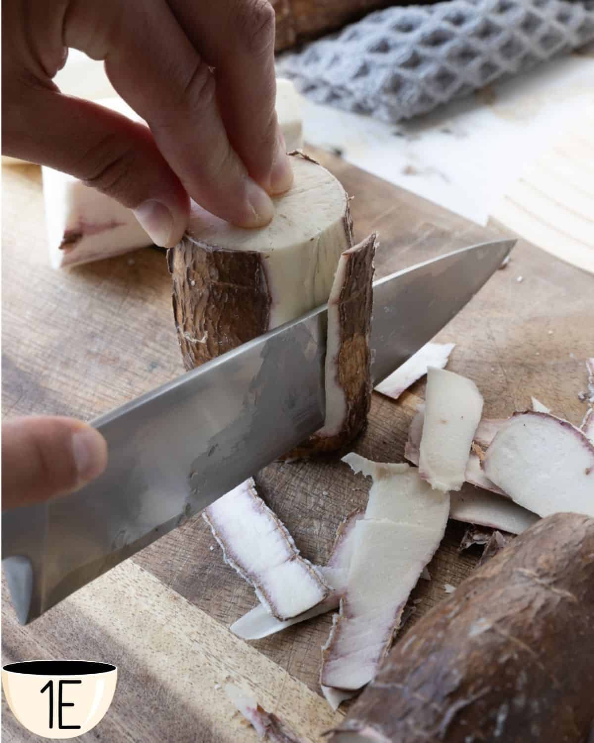 A hand slicing the peeled yuca root into fry-sized pieces on a wooden cutting board.