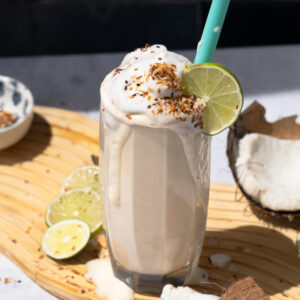 Creamy coconut shake with whipped topping, toasted flakes, and lime, styled with coconut shell and slices on a wooden board.