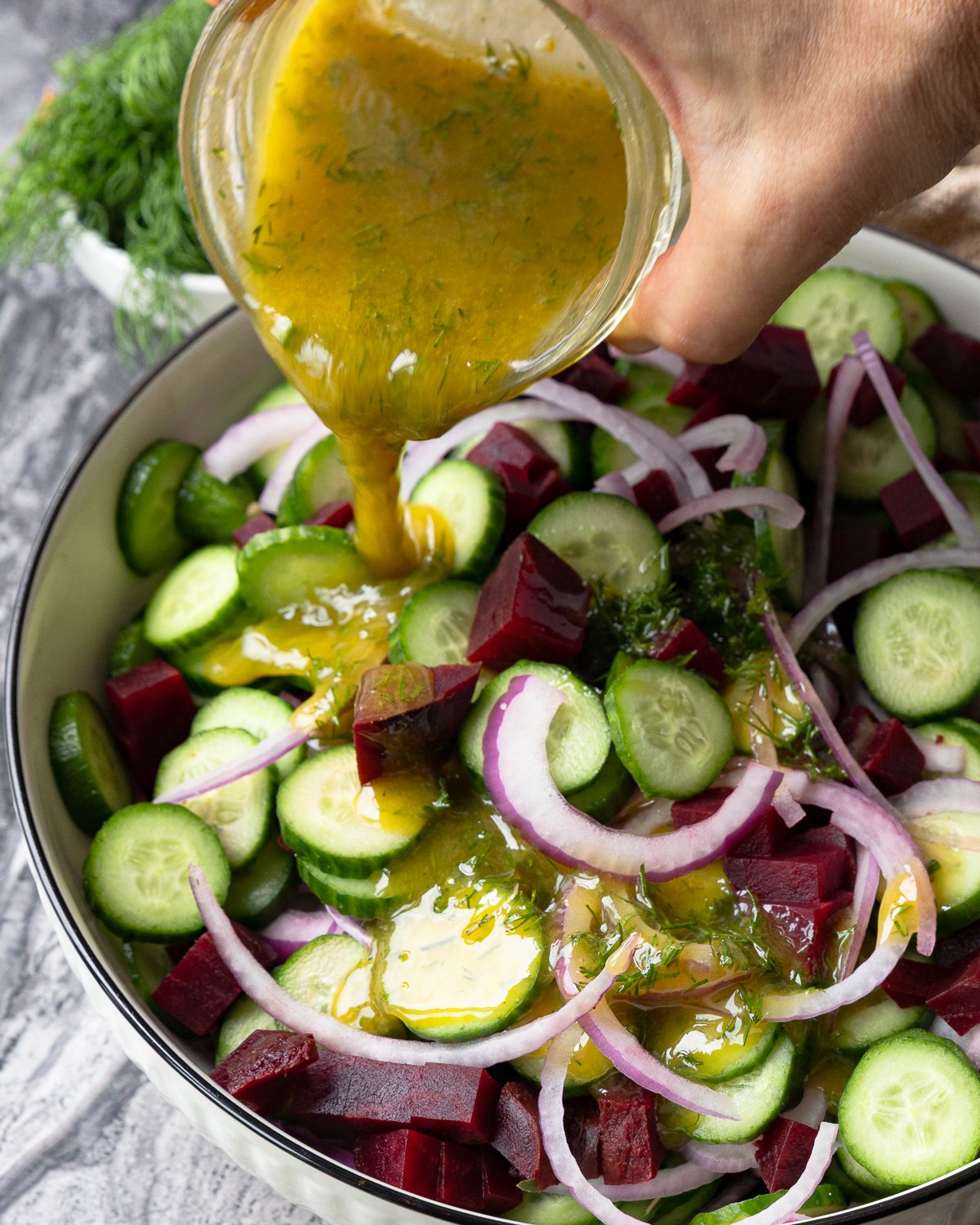 Pouring lemon-mustard dressing over sliced cucumbers, beets, and red onion in a large salad bowl.